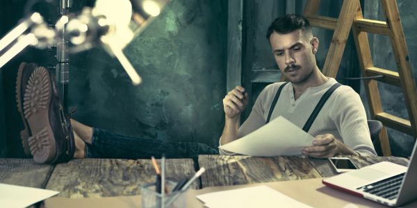 Man with suspenders reading paper at rustic desk.