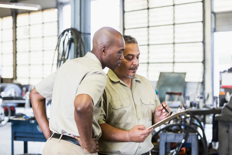 Two multi-ethnic men working for a trucking company, standing in a garage, looking down at a clipboard. The African-American man is in his 30s. His coworker or manager is a mature Hispanic man is in his 40s.