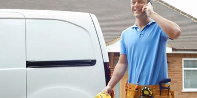 Smiling handyman talking on phone beside a white van with tools in belt.