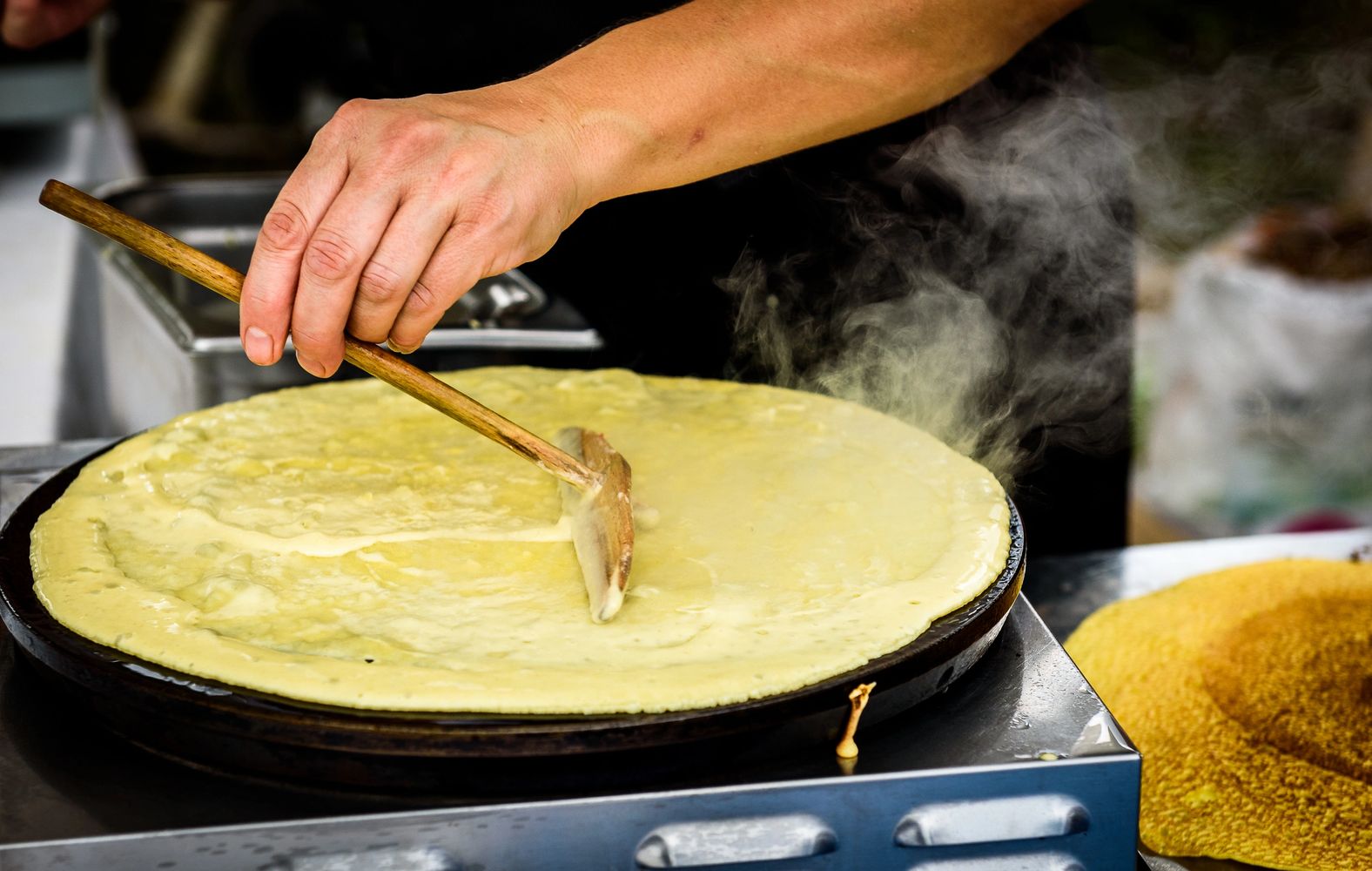A person spreads batter on a hot griddle to make a crepe.