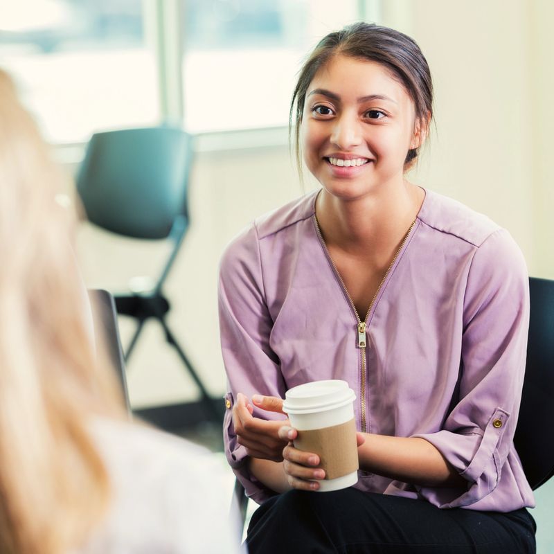 Caring young Hispanic female therapist attentively listens to unrecognizable patient during a session.