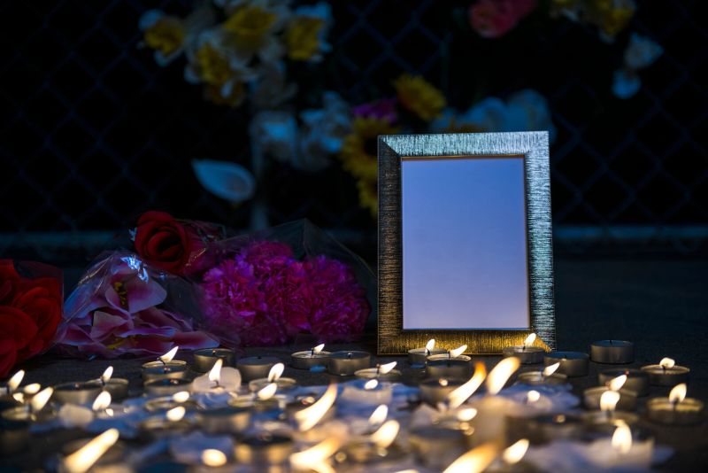 A growing makeshift vigil for victims made out of various size candles on a concrete sidewalk at night with and empty gold picture frame, flowers and some flowers stuck in the chainlink fence in the background. These types of memorial vigils happen after a tragic event such as a mass shootings, terrorists acts or accidents.