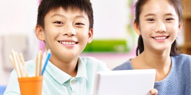 Two smiling children with a tablet and pencils in a bright room.