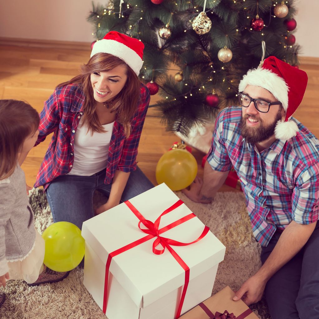 Family in Santa hats exchanging gifts by decorated Christmas tree.