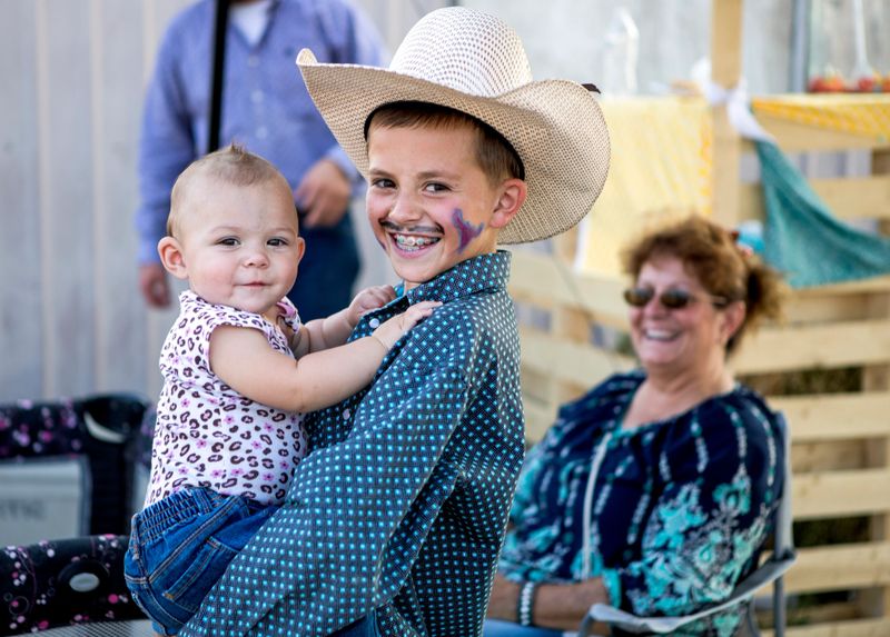 A young rodeo cowboy holds his baby sister at the rodeo as their grandmother looks on.