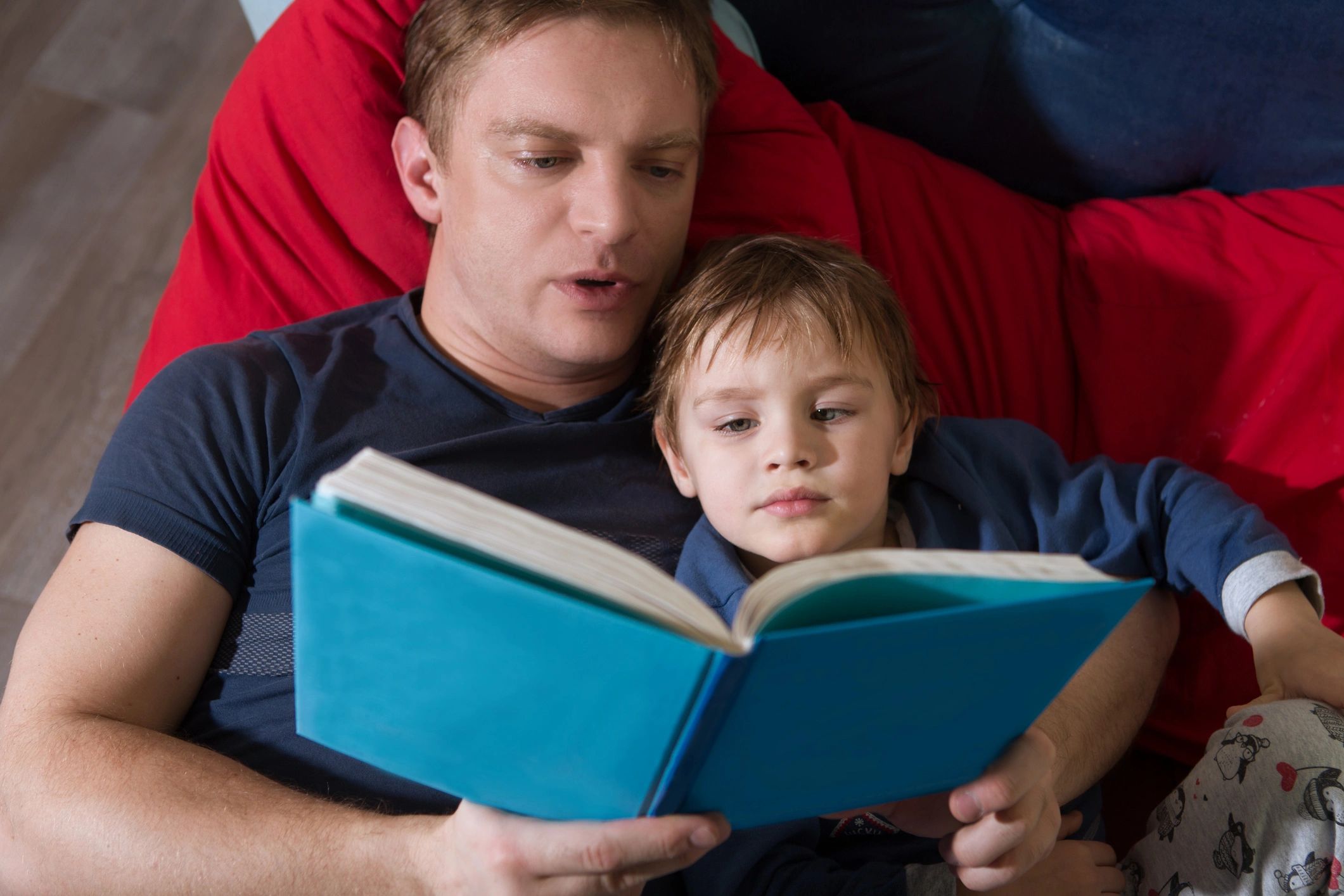 A father reading a book to his son on a red bean bag.