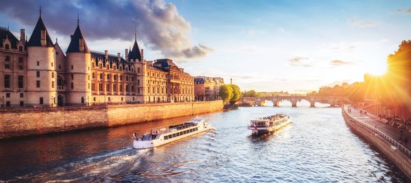 Boats sail on a river near historic buildings at sunset.