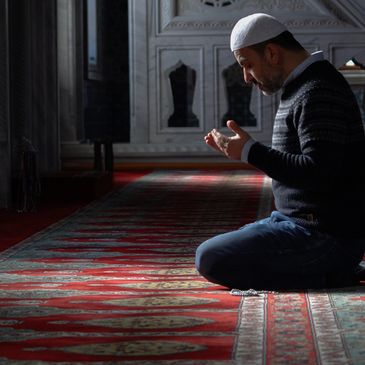 A man kneeling and praying inside a mosque with intricate carpet and architecture.