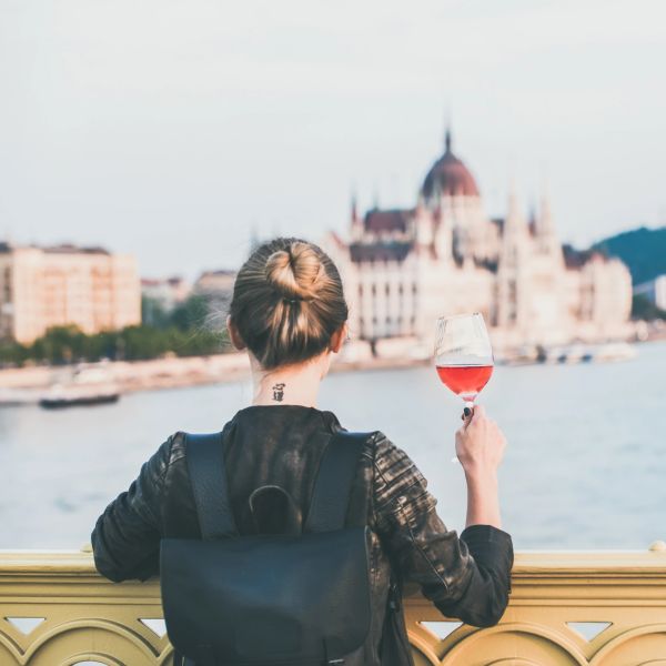 Person with a backpack holding a glass of rosé wine overlooking a river and historic building.