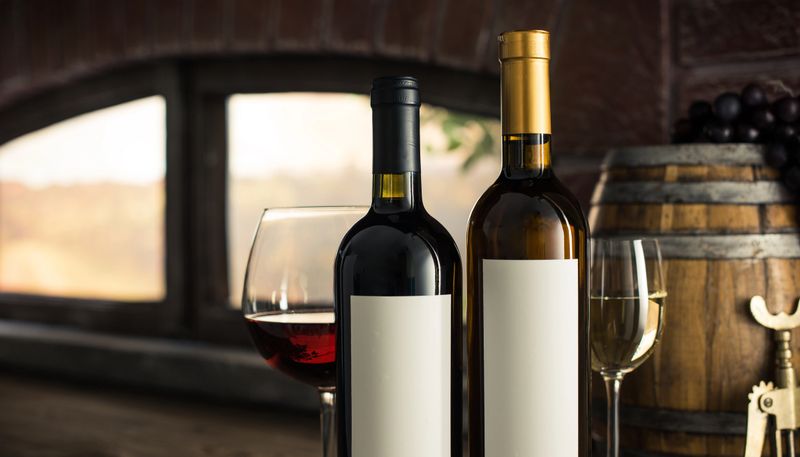 Wine bottles with blank label, wine glasses and barrel next to the wine cellar window; country vineyard view at sunset on the background