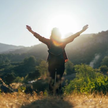Person standing with arms raised facing the sun in nature.
