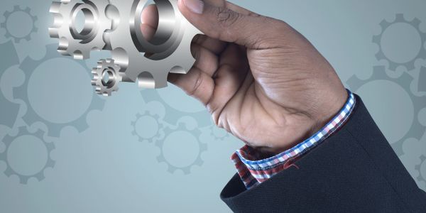 Hand holding a silver gear with interconnected gears in the background.