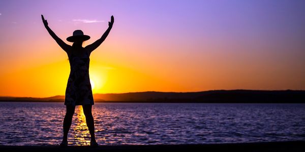 Silhouette of a person with arms raised at sunset by the water.