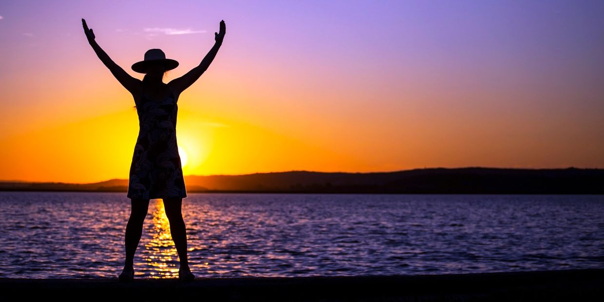 Silhouette of a person with arms raised at sunset by the water.