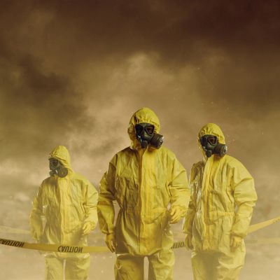 Three people in yellow hazmat suits and gas masks amid smoky surroundings.