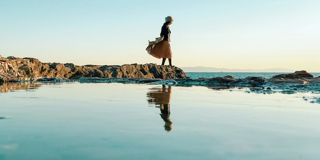 A person stands on a rocky shore with their reflection in the calm water.