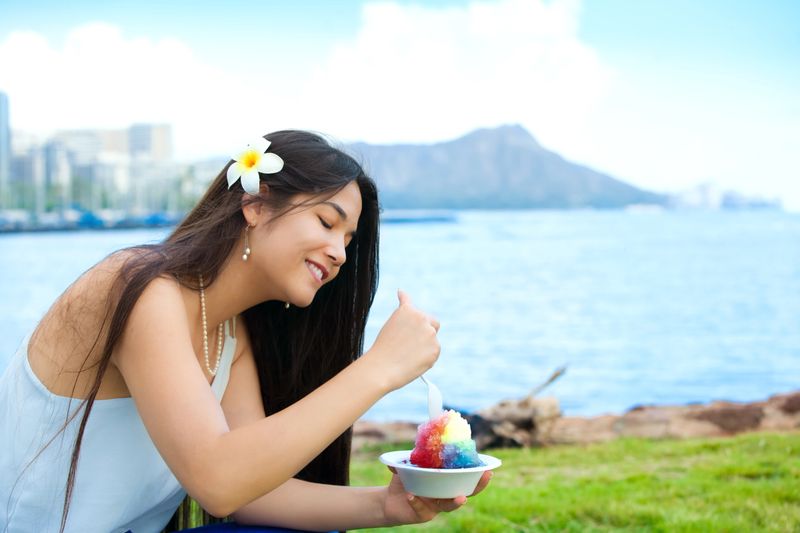 Smiling biracial Thai, Caucasian teen or young woman at Magic Lagoon eating shave ice. Waikiki and Diamond Head in background.