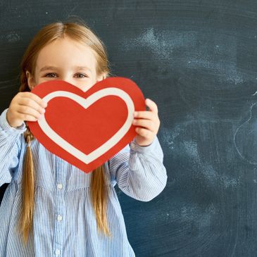 Girl holding red heart cutout in front of chalkboard.