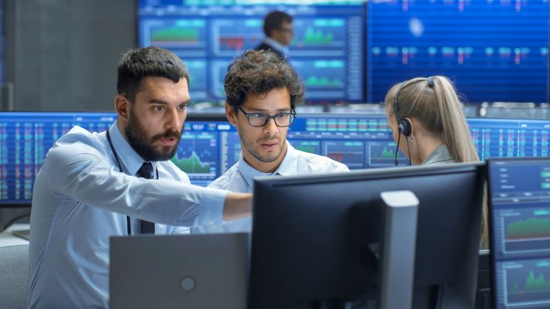Professional Broker Consults Stock Exchange Trader at His Workstation. Multi-Ethnic Team at Stock Exchange Office is Busy Selling and Buying Stocks on the Market. Displays Show Relevant Data Numbers.