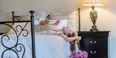 Woman lying on bed smiling, holding a pink flower, with a vase of flowers and lamp on nightstand.