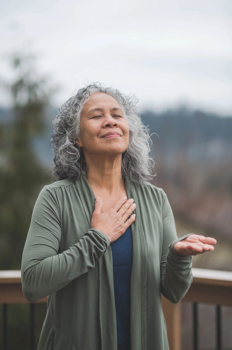 Mature and vibrant Hawaiian woman does yoga outside on an early morning.  One hand is on her chest and the other is facing upwards, palm open. Her face is lifted up and her eyes are closed.
