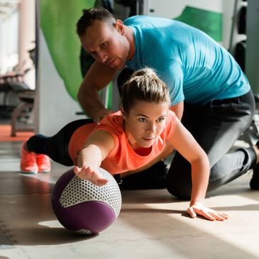 Trainer assists woman stretching with medicine ball at gym.