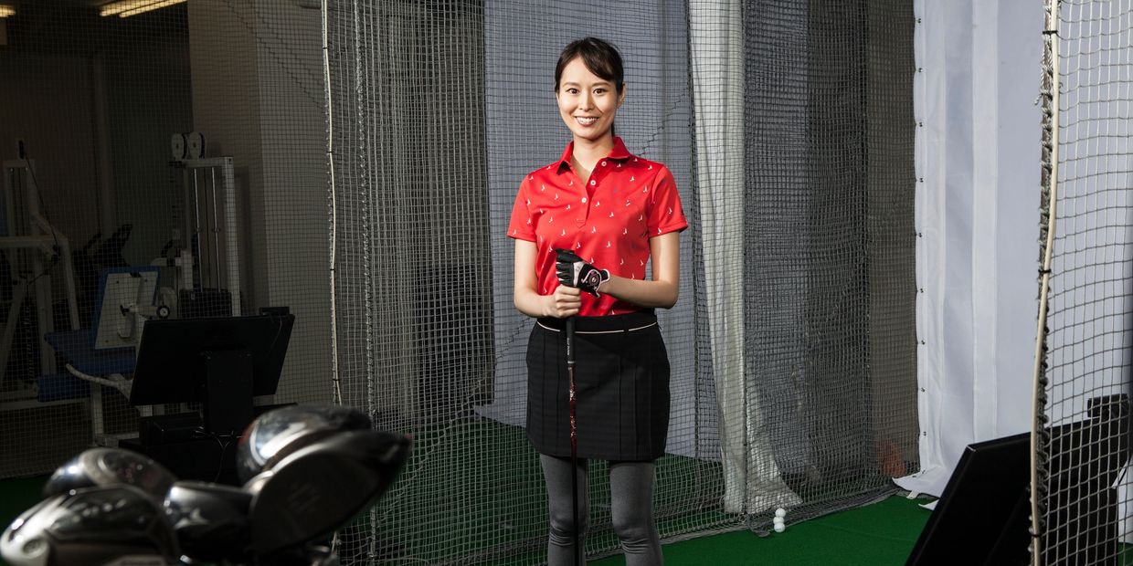 Smiling woman in golf attire holding a club at an indoor golf practice area.