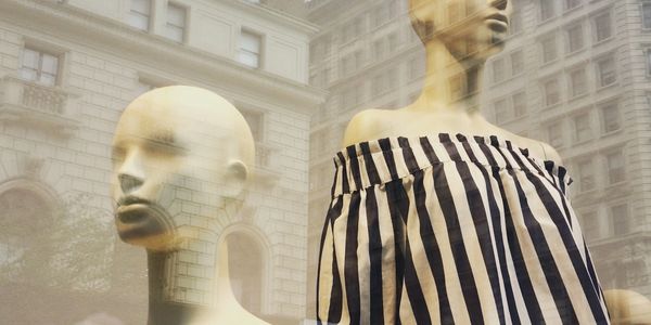 Two mannequins displayed in a shop window with a building reflection.