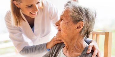 A caregiver warmly supports an elderly woman sitting in a chair.