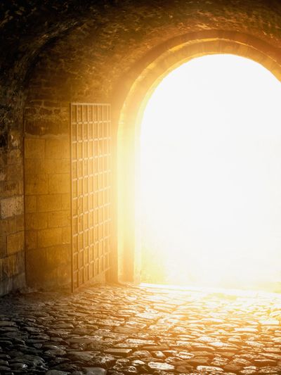 Sunlight floods through an old stone archway with an open iron gate.