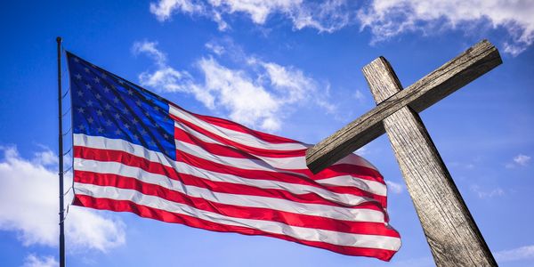 American flag waving next to a wooden cross under a blue sky.