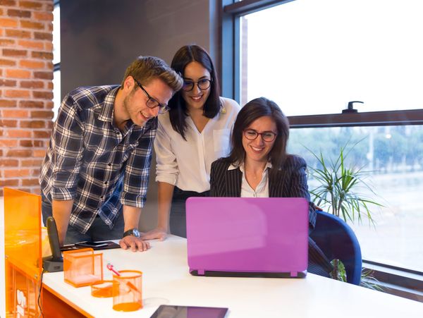 Three colleagues happily collaborate around a laptop in a modern office.