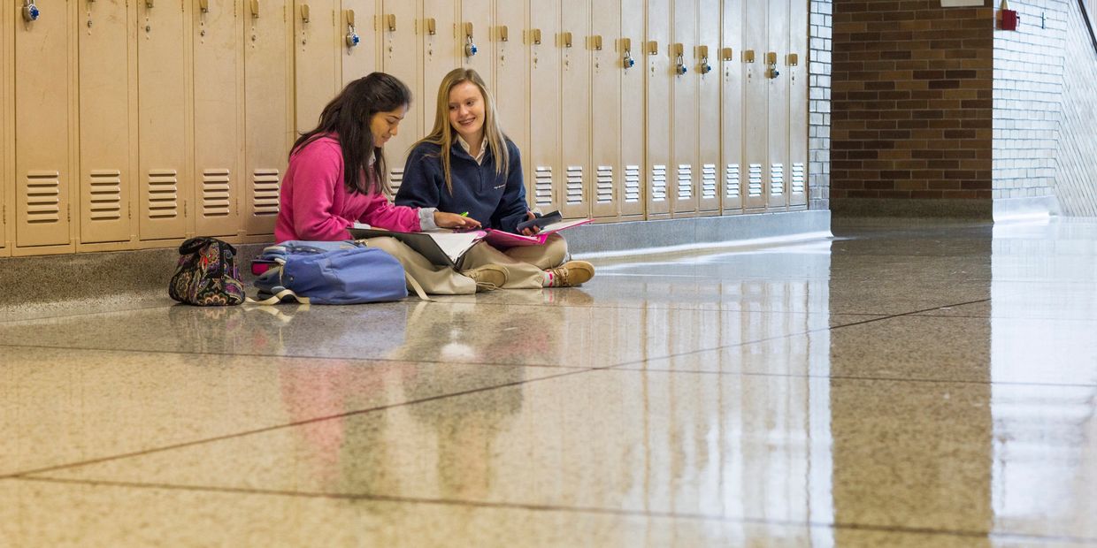 Two students sitting in a hallway by lockers, working together with notebooks.