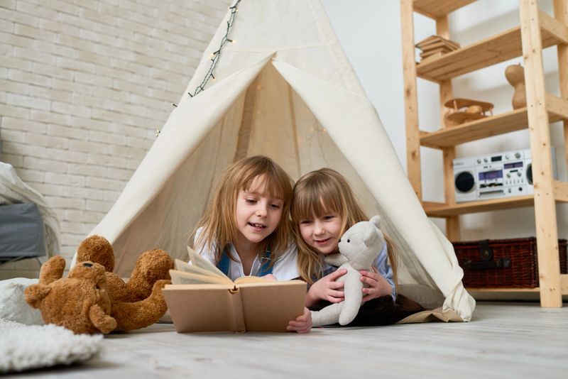 Curious young readers enjoying kids book, elder sister reading fairy tale to young sister while they relaxing in teepee at home