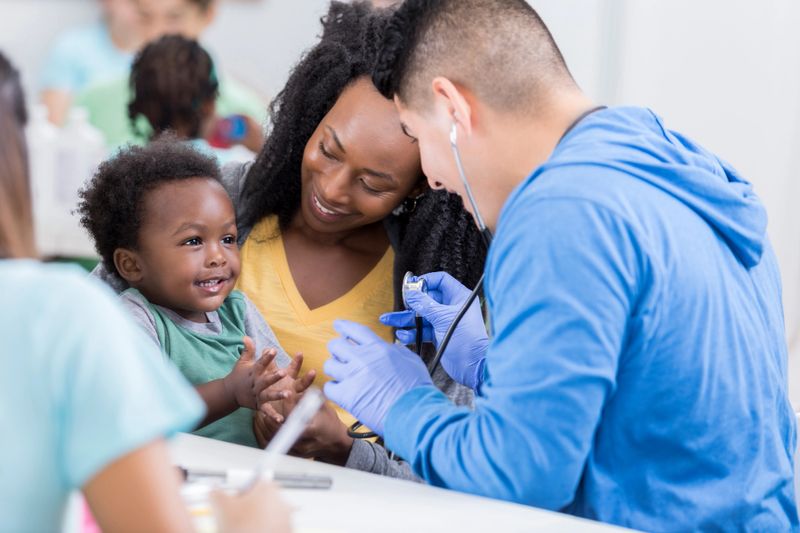 An adorable little toddler boy sits in his mother's lap in a volunteer run medical clinic and claps for a male nurse. He is unrecognizable as she leans in to listen to his heart.