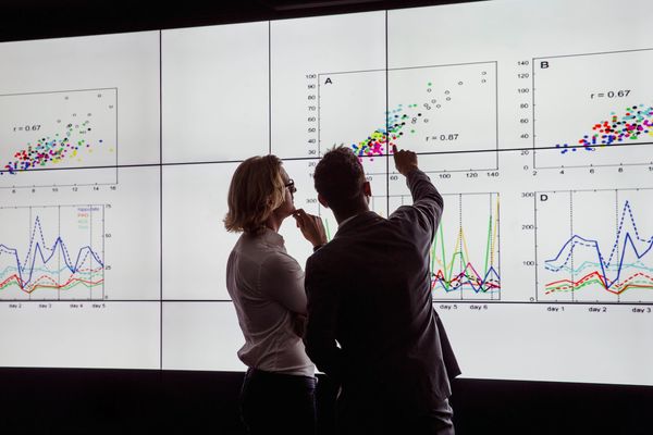 Two professional colleagues analyze and discuss data presented on a large, multi-screen display.