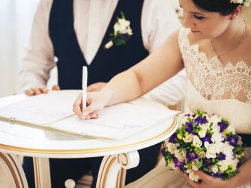 Bride signing a document while holding a bouquet, groom beside her.