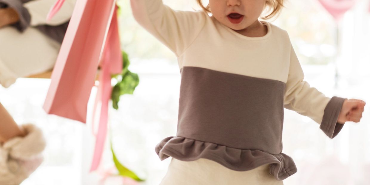 A child in a cream and gray dress holding a pink bag, mid-motion indoors.