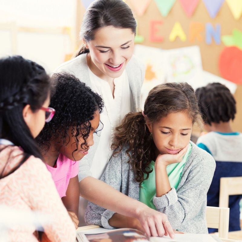 Caring young female kindergarten teacher shows schoolgirls something in a book. The girls are looking at the book.