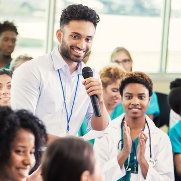 A man speaks into a microphone while medical professionals listen and applaud.