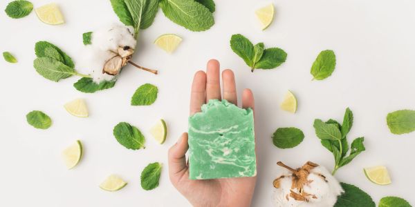 Hand holding green soap surrounded by mint leaves and lemon slices.