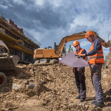Two construction workers review plans at a busy construction site with heavy machinery.