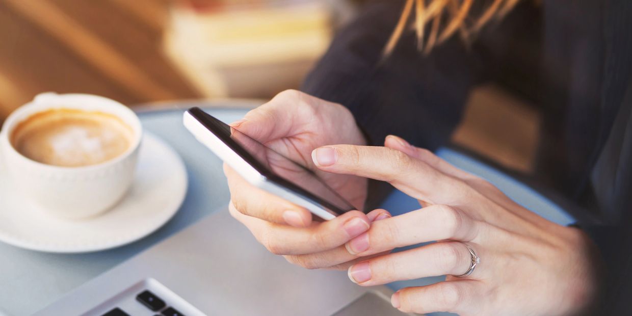 Person using smartphone at a table with a laptop and coffee.