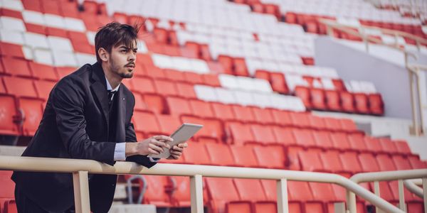 Man in a suit holding a tablet, standing in an empty stadium.