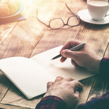 Image of someone writing in a journal with glasses and coffee in the background