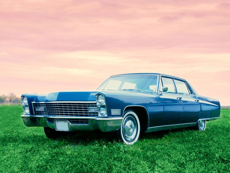 Parked in a field at sunset, with green grass and a purple pink sky wrapping the Fleetwood Cadillac 1967 vehicle in an evening glow.  An old car with its manufacturer’s blue paint, pimped up to drive and look like new.  View of the front grill with lights on.