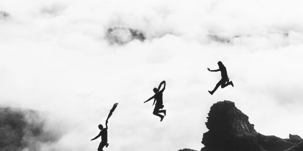 Silhouetted person jumping between rocks above clouds in black and white.