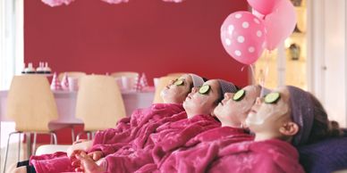 Four girls relaxing with facial masks and cucumber slices on eyes in pink robes at a party.
