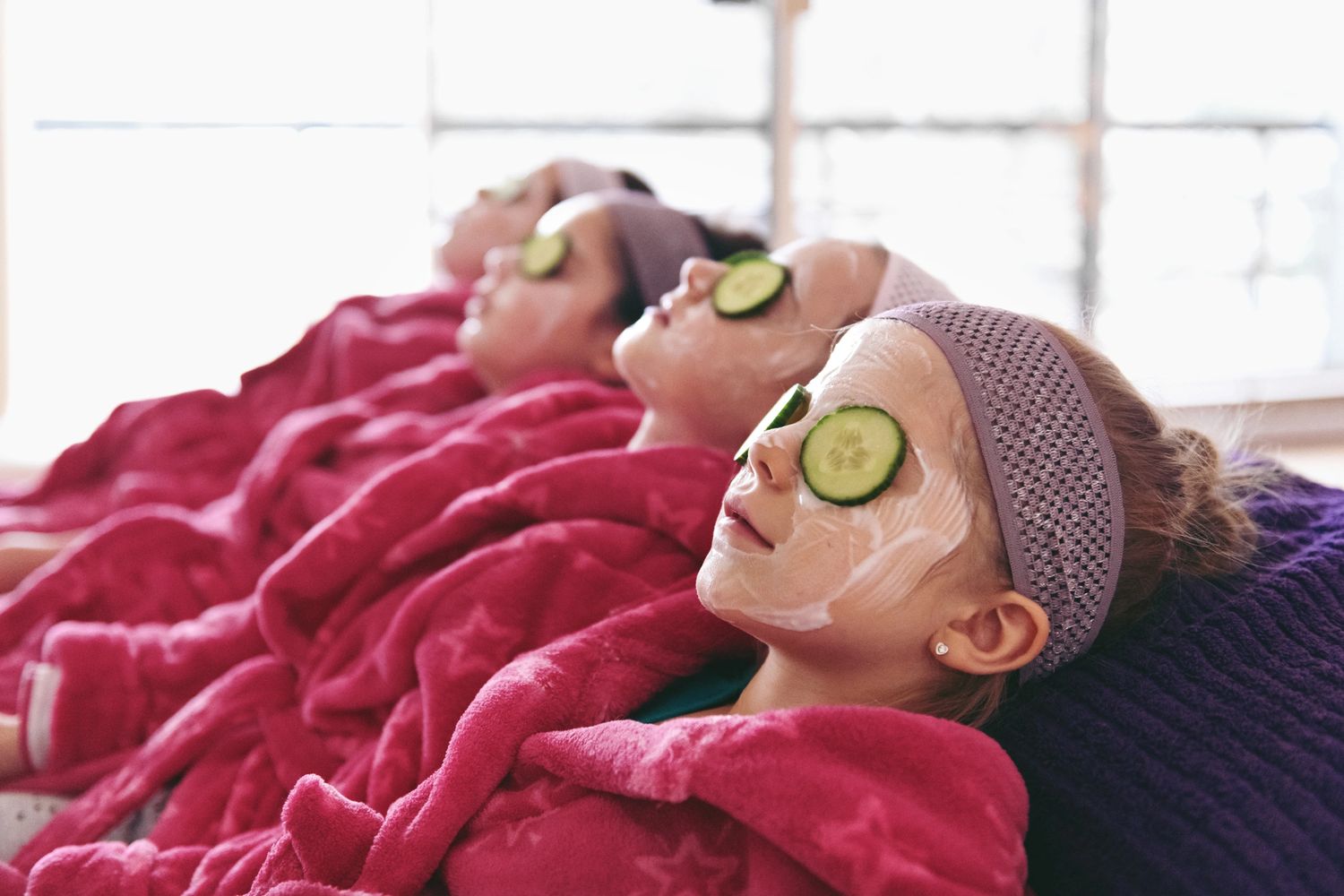 Four girls relaxing with facial masks, cucumber slices on eyes, and pink robes.