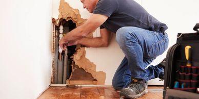 Man fixing pipes behind damaged wall.